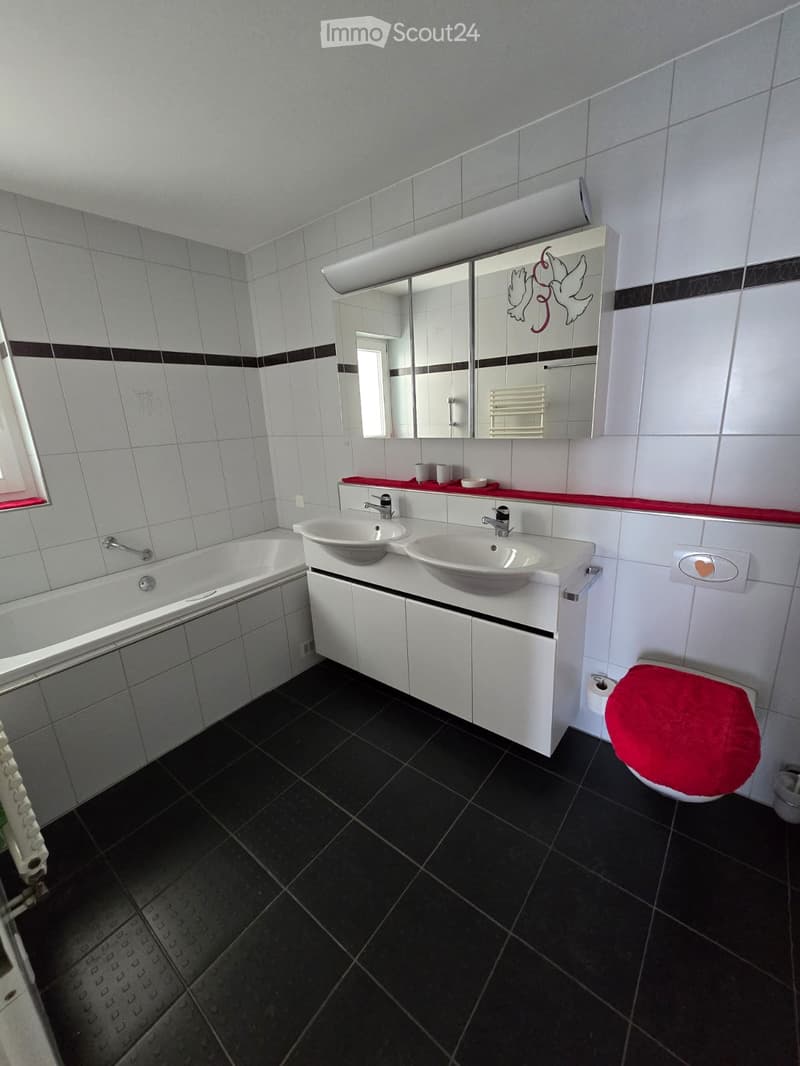 Modern bathroom with black tiled floor, white tiled walls, double sink vanity, bathtub, toilet with red seat cover, and a large mirror with decorative design.