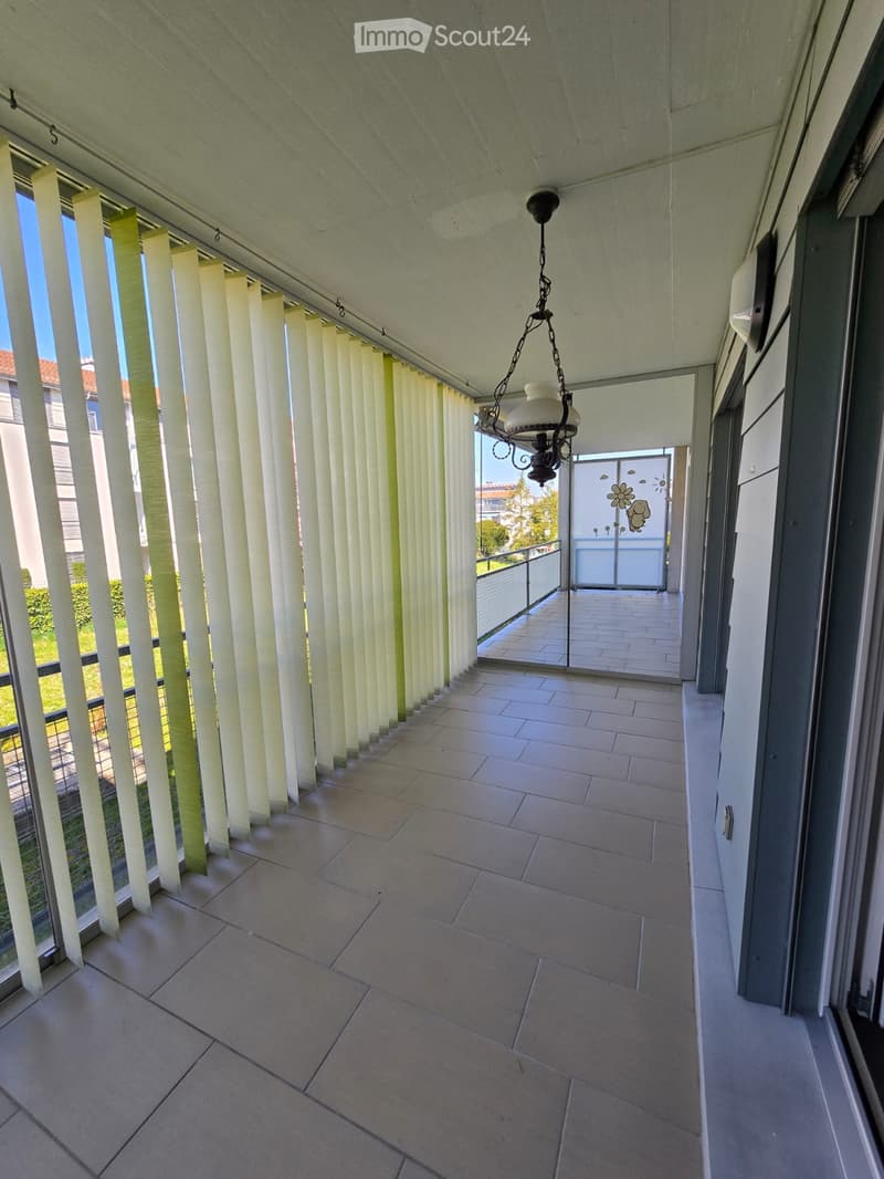 Balcony with tiled floor, vertical blinds, and a hanging light fixture. The balcony has a view of greenery and a neighboring building.