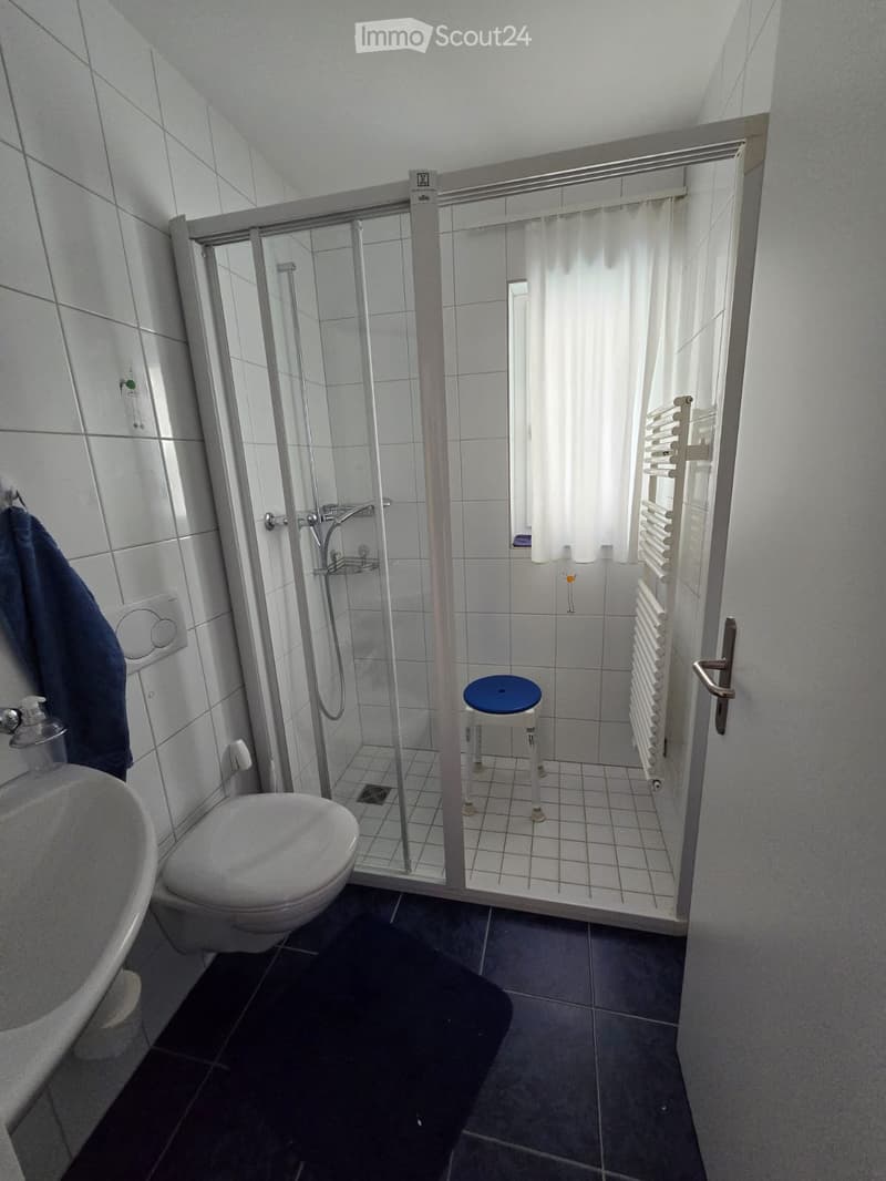 Modern bathroom with white tiles, a glass-enclosed shower, a toilet, a sink, a small blue stool, and a radiator. A towel hangs on the wall, and a window with white curtains allows natural light.