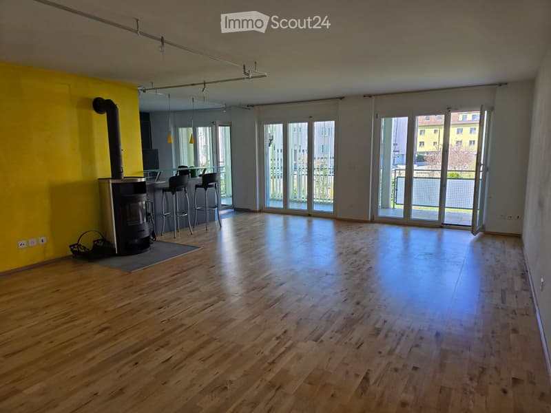 Empty room with wooden flooring, large windows, and a fireplace area with bar stools.