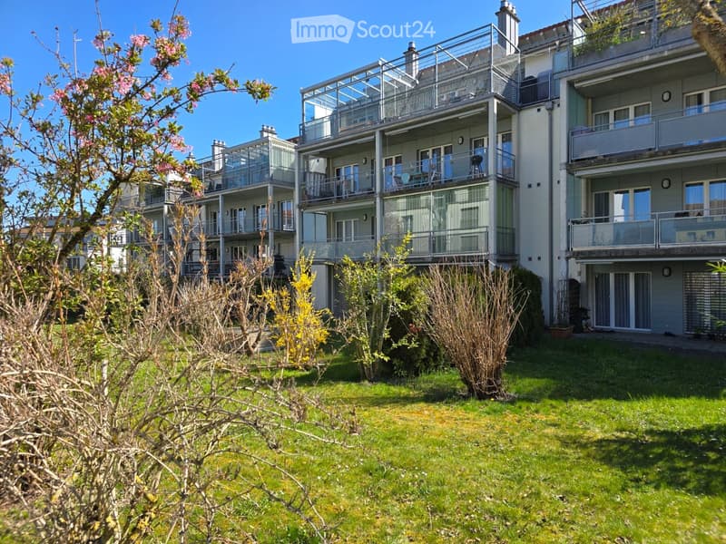 Multi-story residential building with balconies, surrounded by a well-maintained lawn and flowering shrubs.