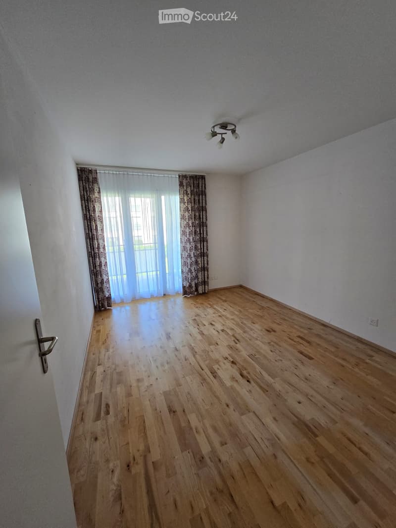 Empty room with wooden flooring, white walls, and large windows covered with sheer curtains. A door is visible on the left side.