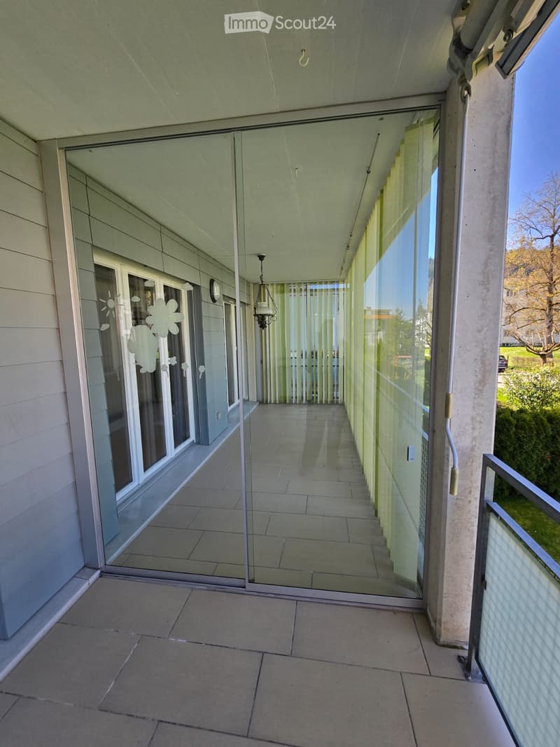 Modern balcony with large glass sliding doors, tiled floor, and a view of the surrounding greenery.