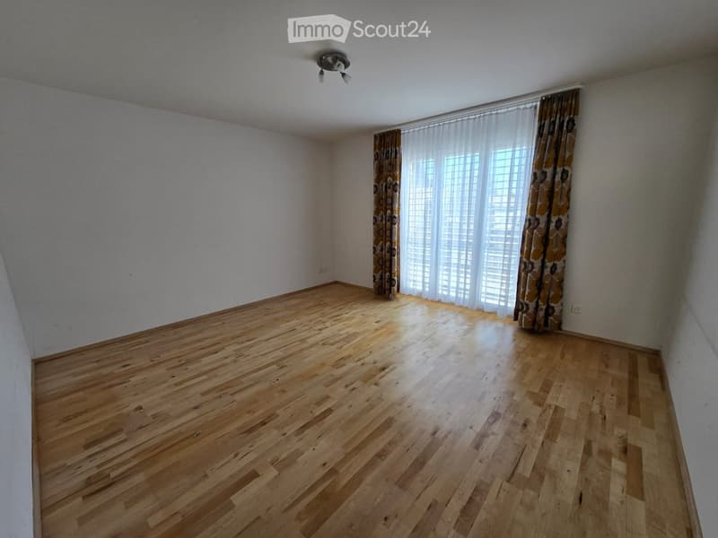 Empty room with wooden flooring, white walls, large window with curtains, and a ceiling light fixture.