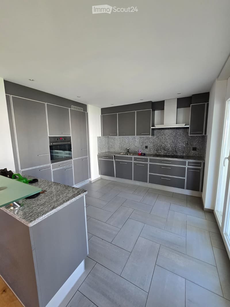 Modern kitchen with grey cabinets, granite countertop, built-in oven, and sink. The space features tiled flooring and ample natural light from a large window on the right.