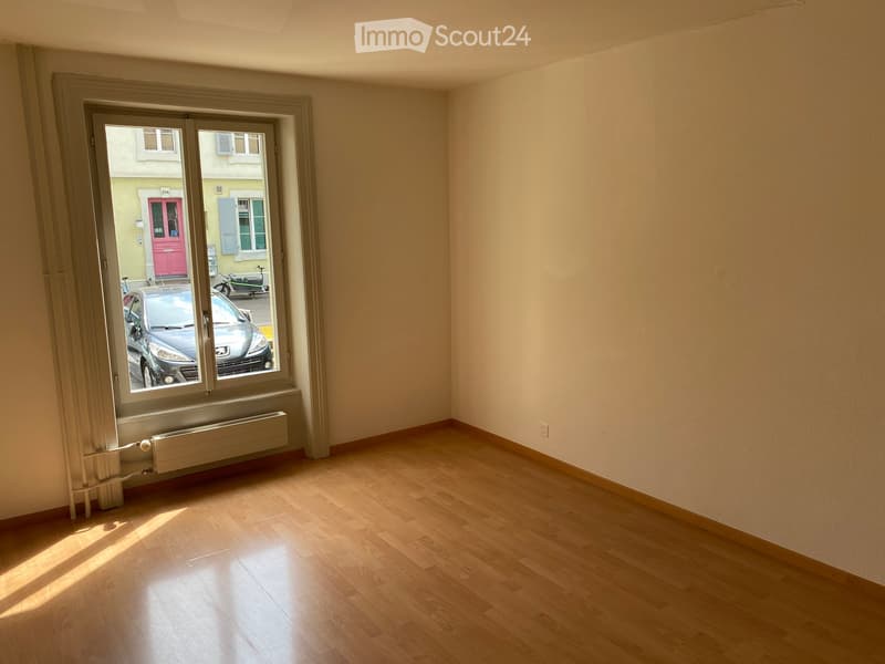 Empty room with wooden flooring, large window with view of street and parked car, radiator below window, light-colored walls.