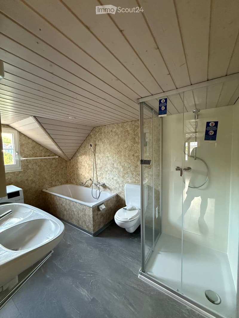 Modern bathroom with a bathtub, toilet, sink, and glass-enclosed shower. Features a sloped wooden ceiling and large window.