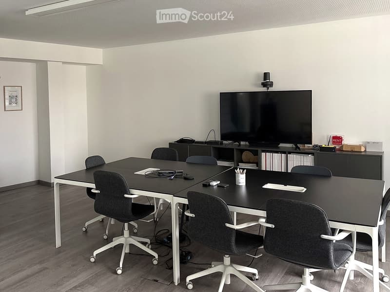 Modern conference room with a large black table, multiple chairs, a large screen mounted on the wall, and a cabinet with books and office supplies.