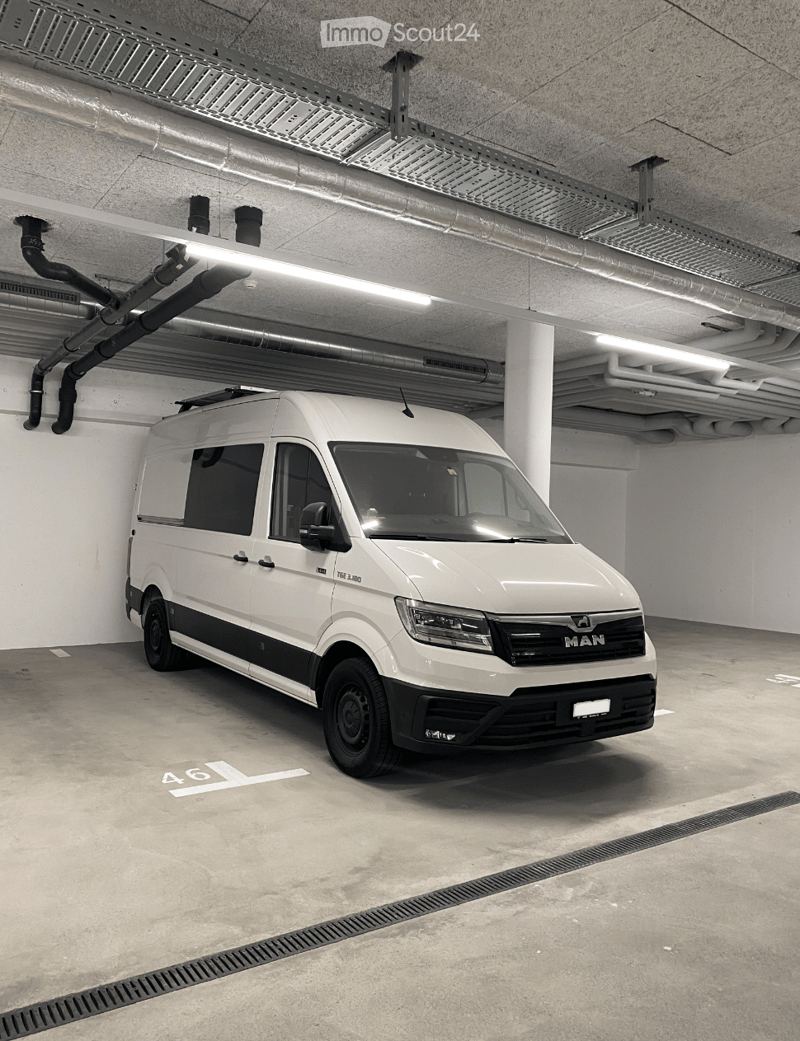 Underground parking garage with a white van parked in one of the designated spots. The area features concrete floors, exposed pipes, and lighting fixtures.