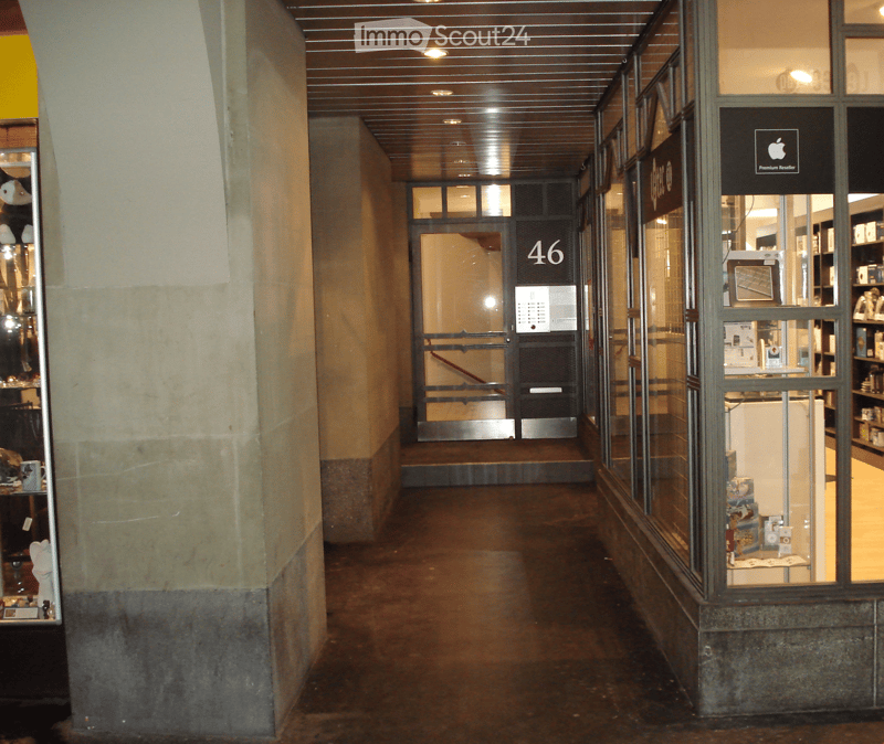 A long, well-lit hallway with a concrete floor and walls, featuring a glass storefront on the right and a door labeled "46" on the left.