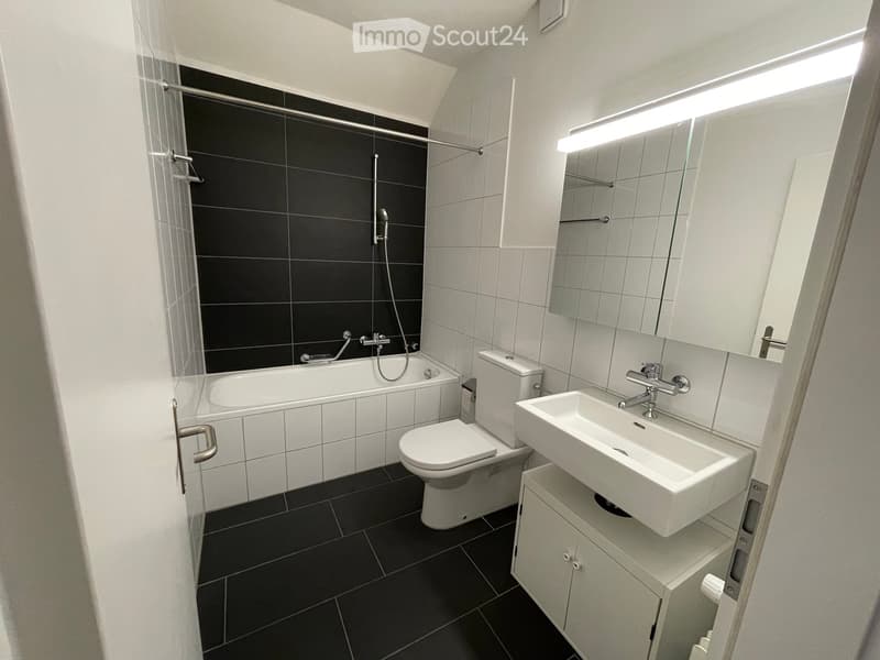 Modern bathroom with white tiles, black accents, bathtub, toilet, sink, and mirrored cabinet.