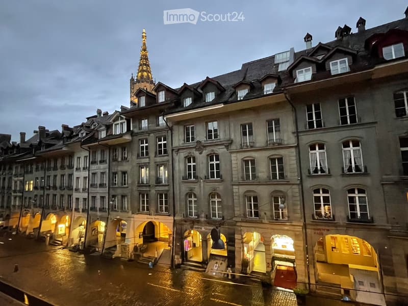 A row of historic European buildings with ornate facades, multiple windows, and illuminated storefronts, with a prominent spire visible in the background.
