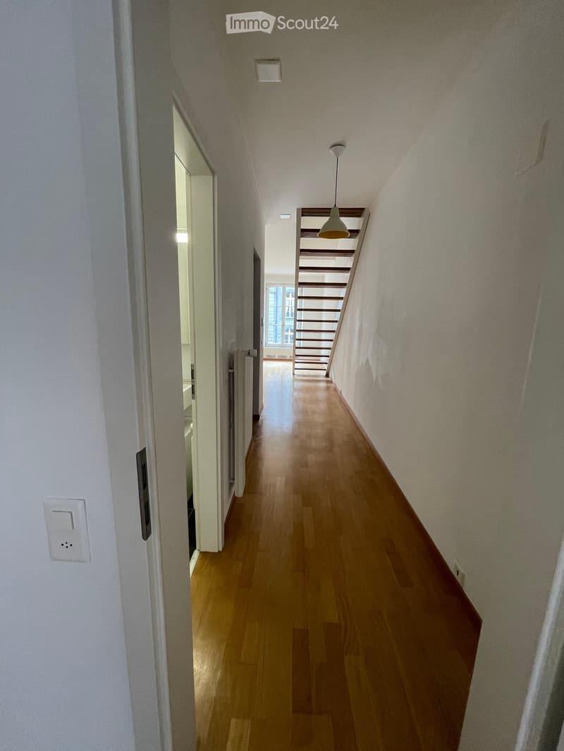 A narrow hallway with wooden flooring, white walls, and a staircase visible at the end. The hallway features a pendant light fixture and a partially open door on the left side.