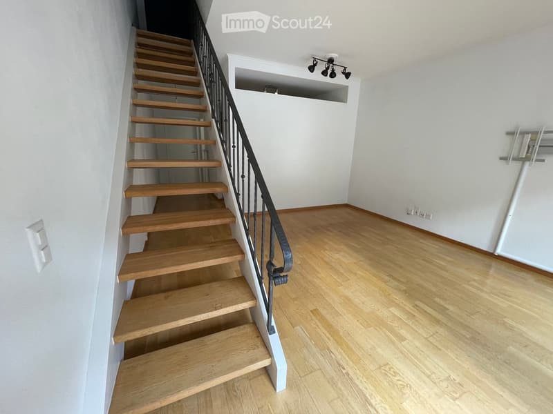 Empty hallway with wooden flooring, white walls, and a wooden staircase with a black metal railing leading upwards.