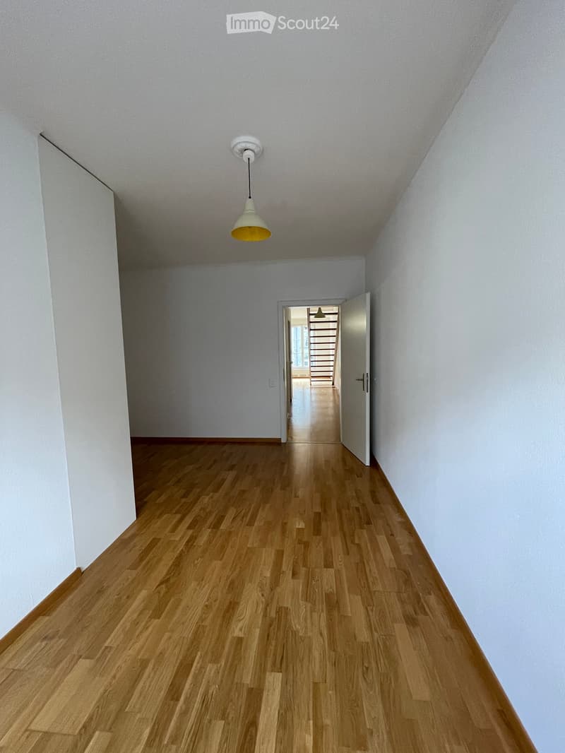 Empty hallway with light wooden flooring, white walls, and a single hanging light fixture. An open door at the end leads to another area.