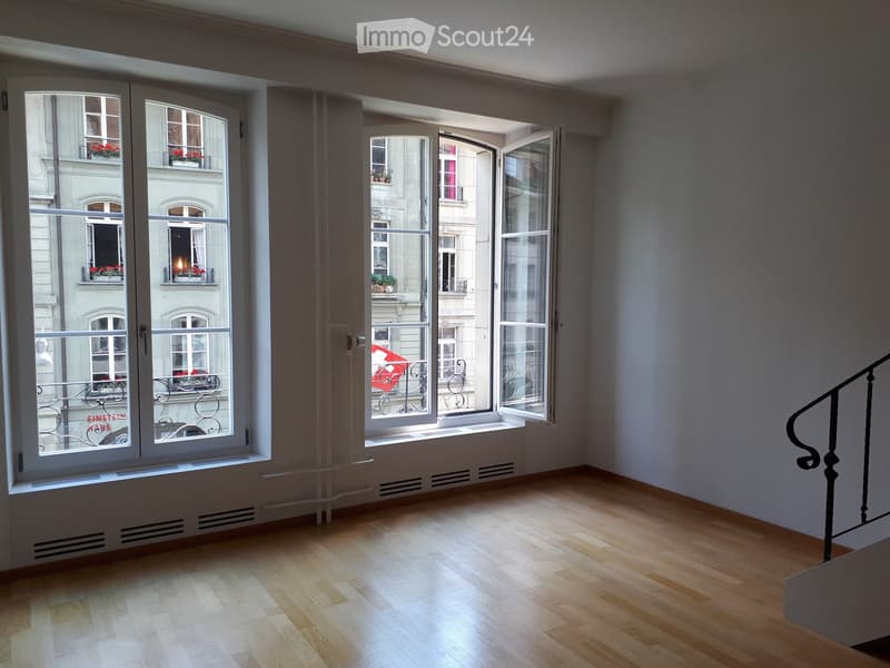 Empty room with wooden flooring, large windows, and a staircase leading upwards. The room has white walls and is well-lit with natural light coming through the windows.