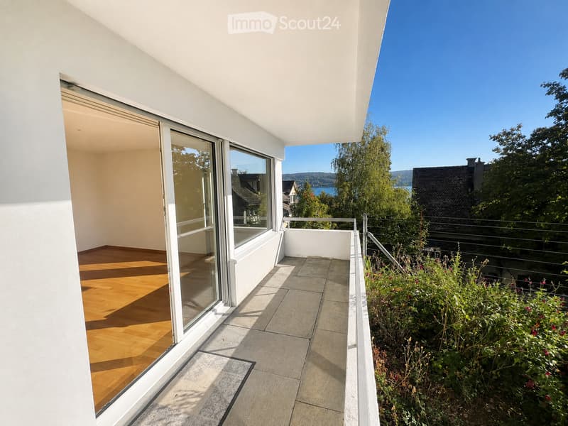Modern balcony with large glass sliding doors, wooden floor inside, tiled balcony floor, metal railing, and scenic view of a lake and mountains.