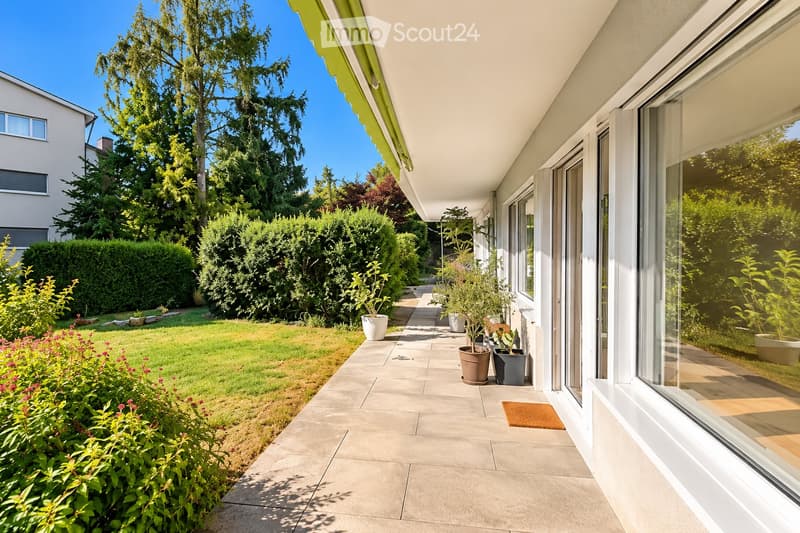 A well-maintained garden with a paved pathway, potted plants, and a neatly trimmed lawn, bordered by a hedge and trees.