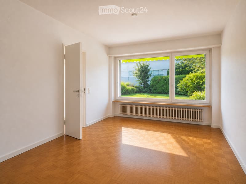 Empty room with wooden flooring, white walls, large windows with a view of greenery, and a radiator below the windows.