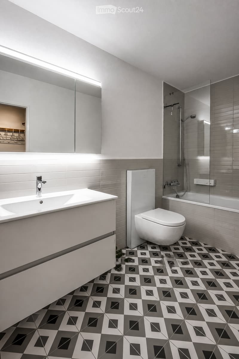 Modern bathroom with white tiles, geometric patterned floor, sink, toilet, bathtub, and shower area.