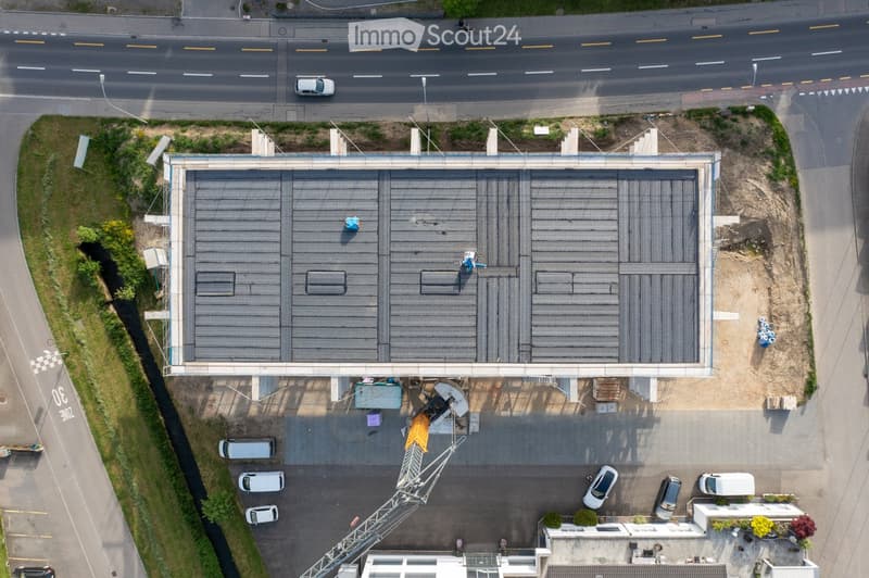 Aerial view of a large industrial building with a flat roof, surrounded by a parking lot and adjacent road. The building has multiple sections and appears to be under construction or maintenance, with visible scaffolding and equipment.