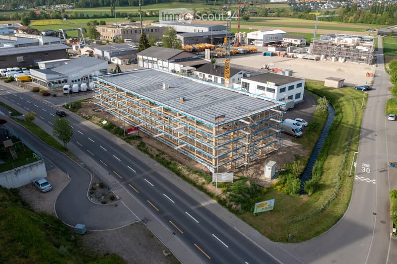 Aerial view of an industrial area with multiple buildings, some under construction, surrounded by roads, vehicles, and green spaces.
