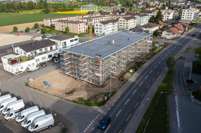 Aerial view of a multi-story building under construction with scaffolding, surrounded by other buildings, parking lots, and a road.
