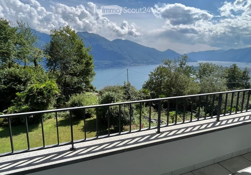 Balcony with metal railing overlooking a scenic lake and mountain landscape, surrounded by greenery.