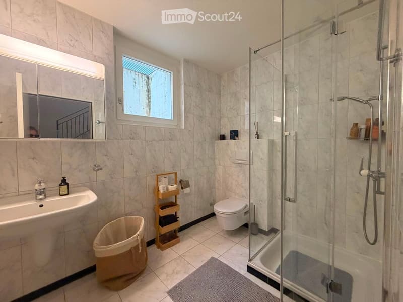 Modern bathroom with marble tiles, a large mirror, a sink, a toilet, a glass-enclosed shower, and a wooden shelf holding toiletries.