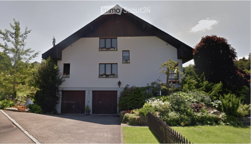 Two-story house with white exterior, dark wooden roof, large windows, and a double garage. The house is surrounded by a well-maintained garden with various plants and flowers.