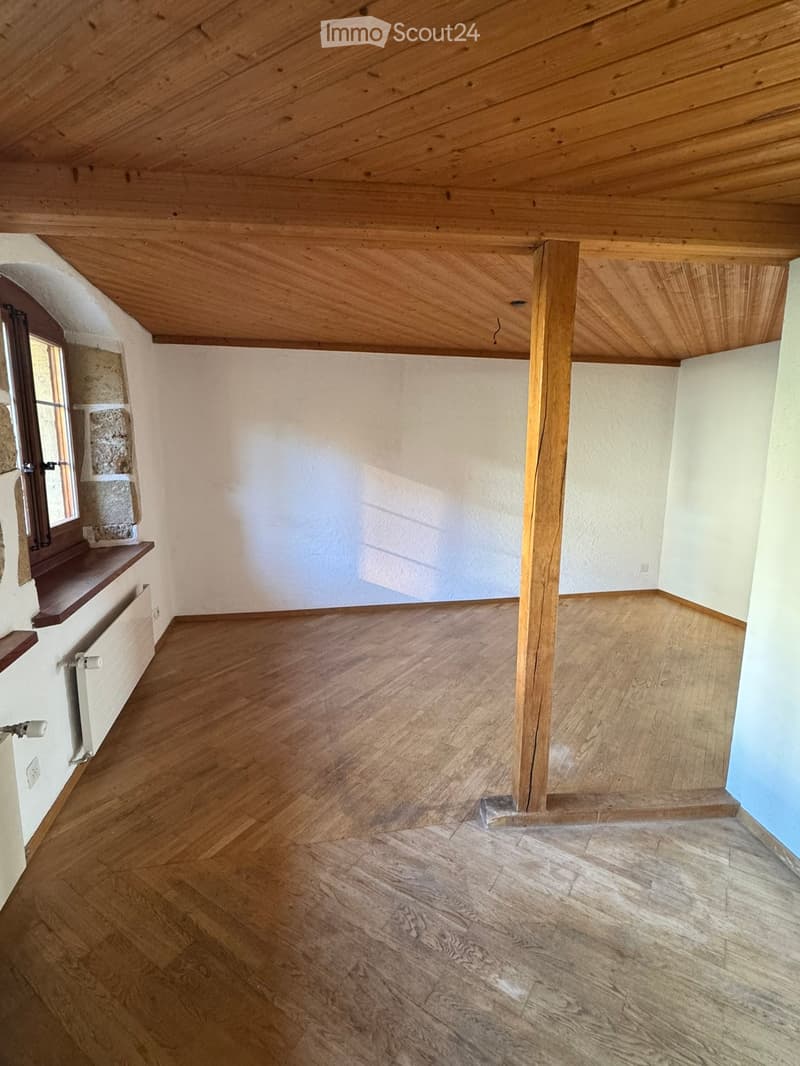 Empty room with wooden flooring, white walls, and a slanted wooden ceiling. Features a window with a wooden sill and a radiator below it.