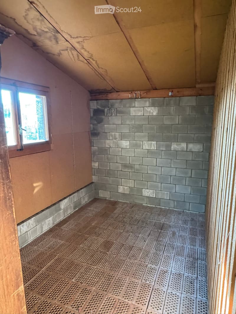 An empty attic space with exposed wooden beams, a slanted ceiling, and a small window letting in natural light. The floor is covered with a perforated metal sheet, and the walls are partially finished with grey tiles and plaster.