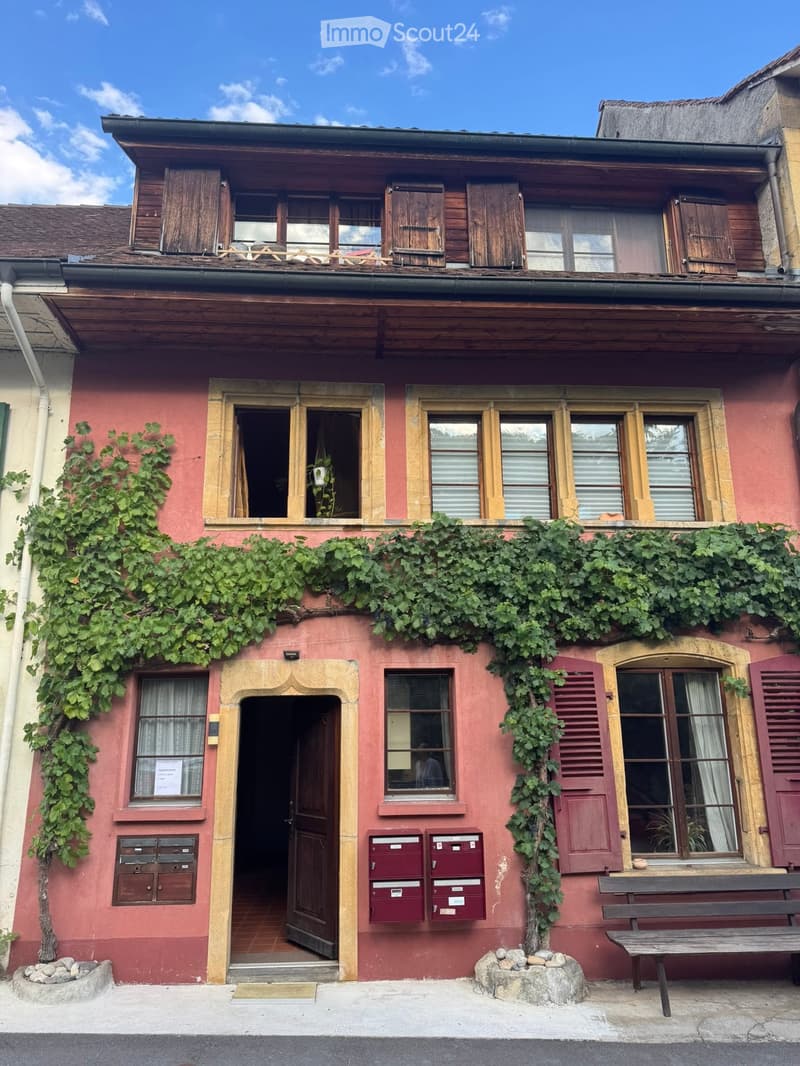 Two-story building with red brick facade, wooden shutters, and ivy-covered walls. Features a central entrance with a small porch, multiple windows, and a wooden bench on the sidewalk.