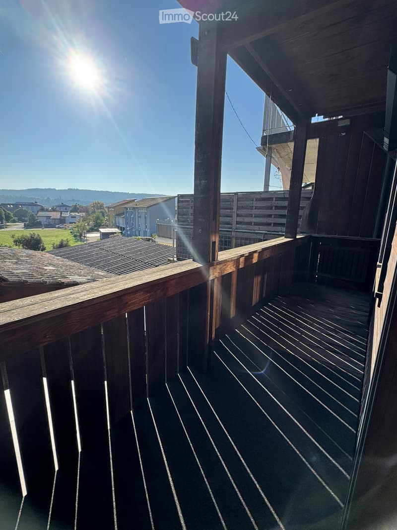 Wooden balcony with railing, overlooking a residential area with houses and greenery.