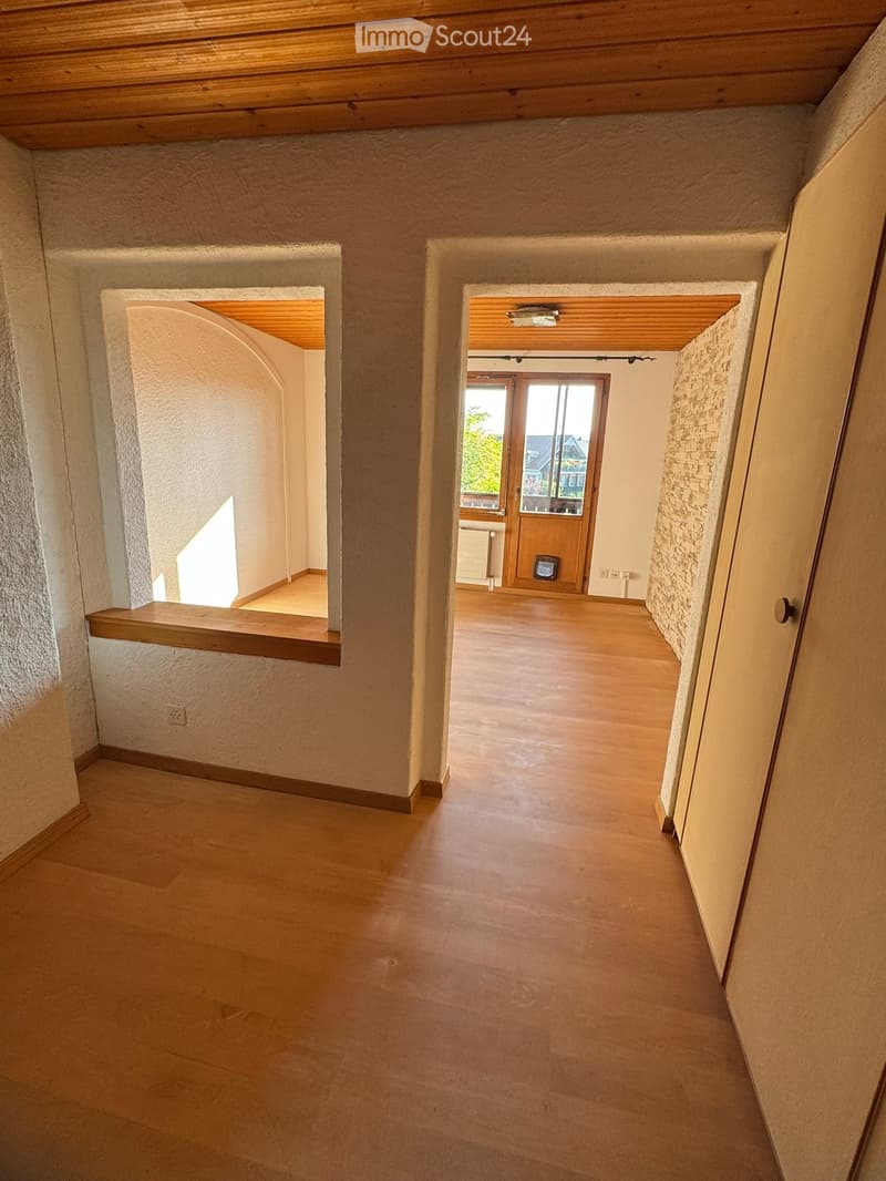 Empty hallway with wooden flooring, textured walls, and arched doorways leading to other rooms. Natural light filters through a small window on the left side.