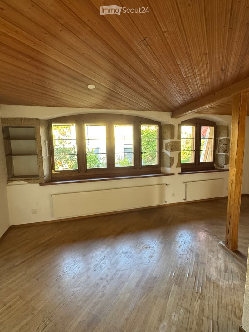 Empty room with wooden floors, wooden slanted ceiling, large windows, and radiators.
