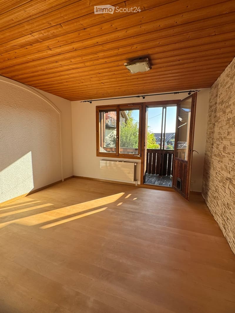 Empty room with wooden flooring, white walls, large windows, and a door leading outside. The ceiling is wooden with built-in lighting.