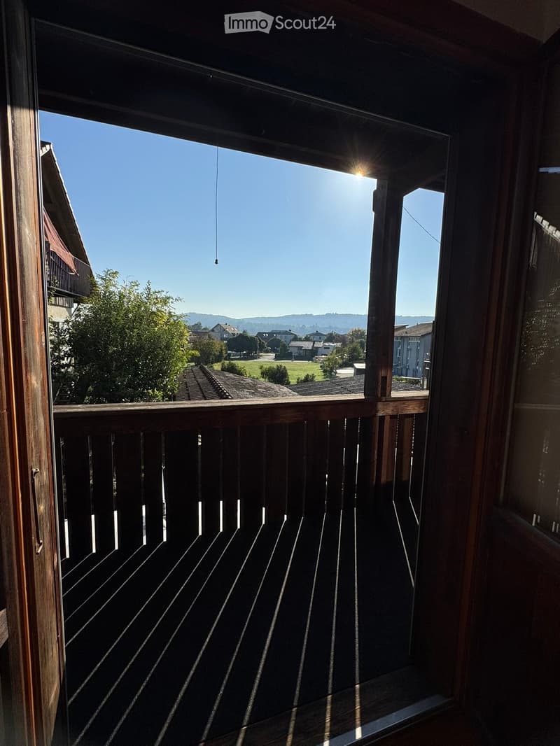 Wooden balcony with a railing, overlooking a residential area with trees and houses under a clear blue sky.