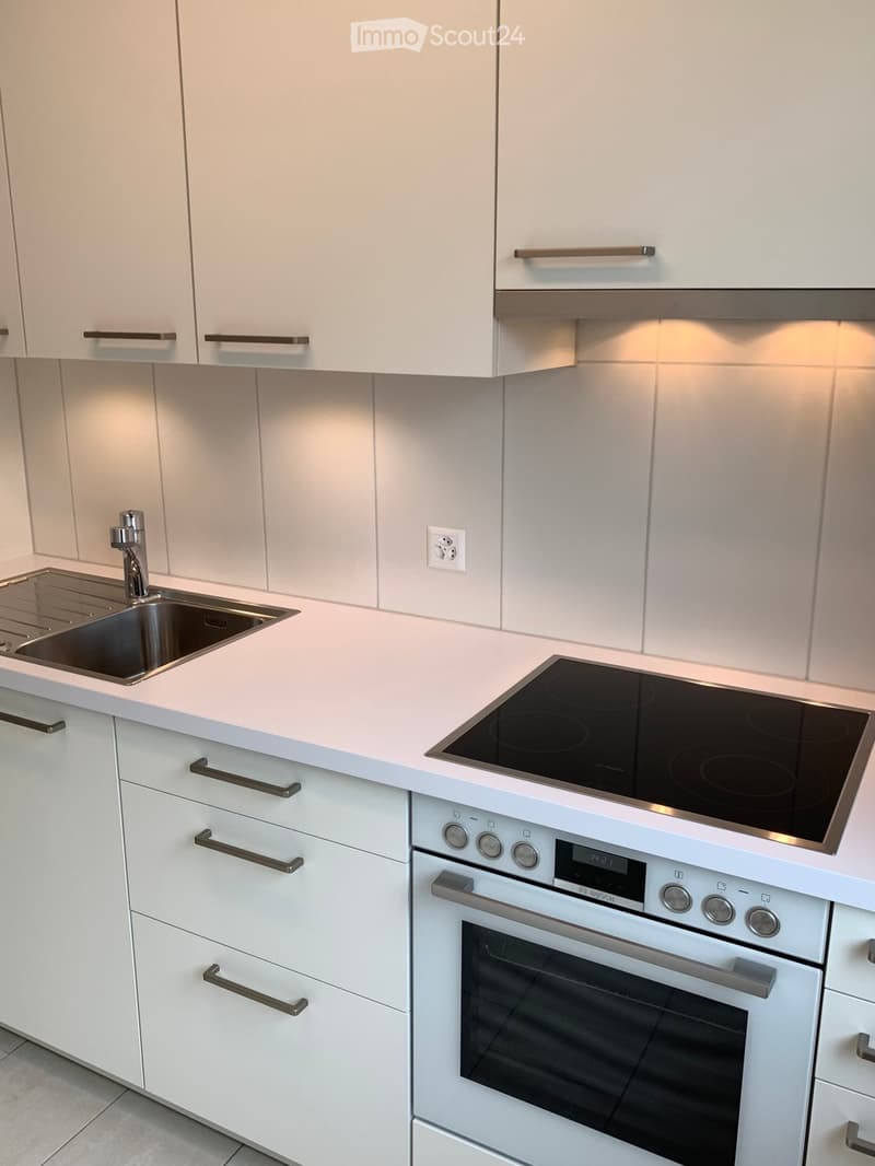Modern kitchen with white cabinets, a stainless steel sink, an induction cooktop, and an oven. The countertops are light-colored, and the backsplash is tiled.