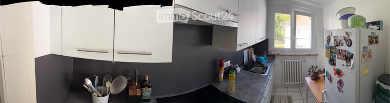 Modern kitchen with white cabinets, stainless steel appliances, and a grey countertop. Visible items include a refrigerator with photos, a sink, a bottle, a cup, a bowl, a cutting board, a spoon, a frying pan, and a small table.