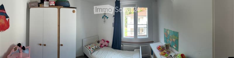 A child's bedroom with a single bed, a wardrobe, a small table with toys, a world map on the wall, and a window with blue curtains.