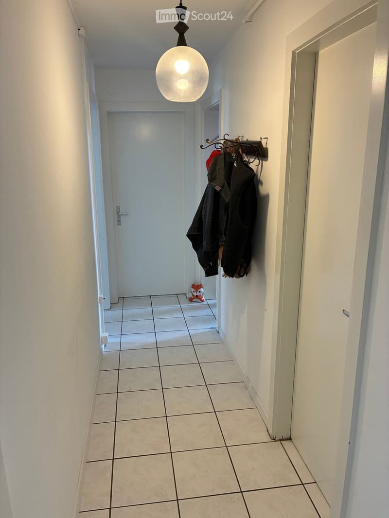 A narrow hallway with tiled flooring, white walls, and a modern pendant light fixture. A coat rack with jackets and a small decorative fox figurine are visible.