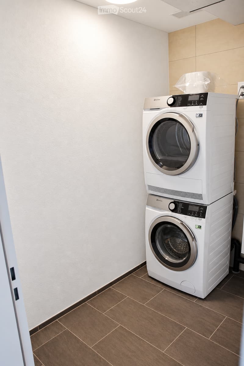 Modern laundry room with a stacked washer and dryer unit, wooden flooring, and white walls.