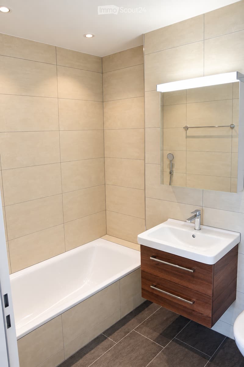 Modern bathroom with beige tiled walls, a white bathtub, a wooden vanity with a sink, and a large mirror with built-in lighting.