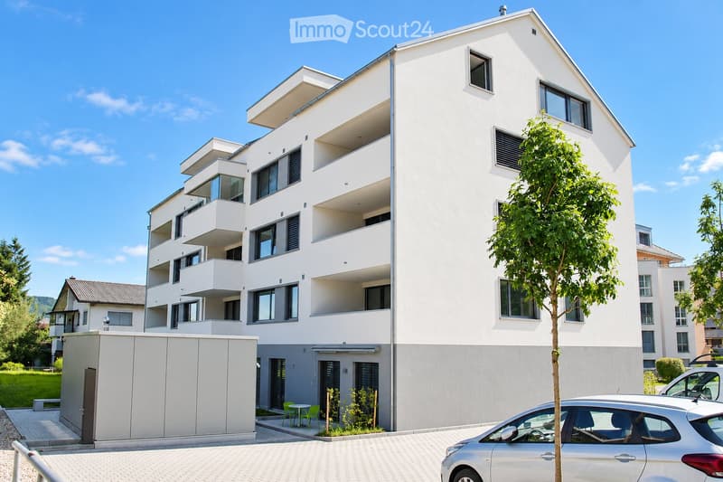 Modern multi-story residential building with white exterior, multiple balconies, and contemporary design. Features include large windows, a small garden area with chairs and a table, and a parking area with cars.