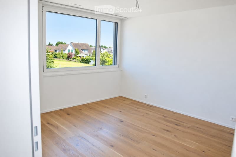 Empty room with light wooden flooring and white walls, featuring large windows with a view of a suburban neighborhood.