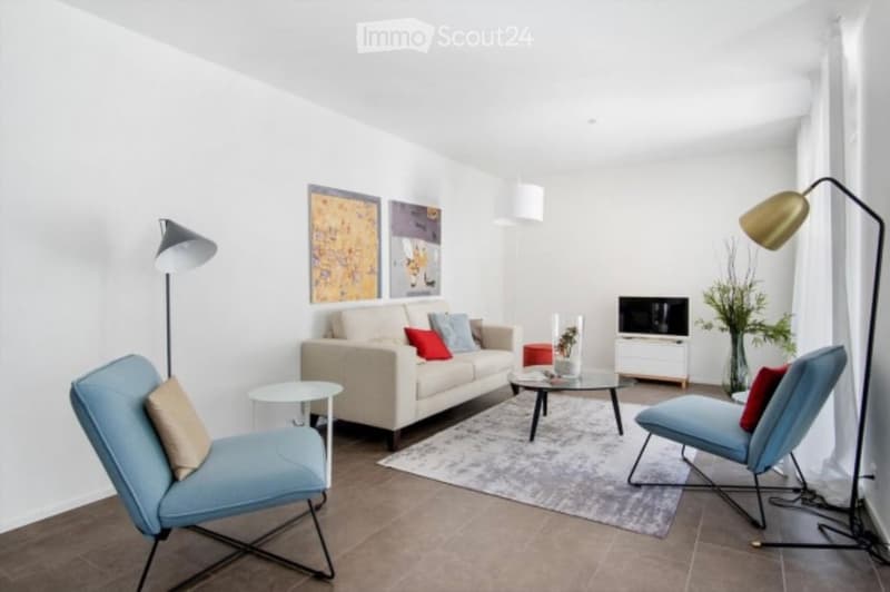 Modern living room with a light-colored sofa, two blue armchairs, a coffee table, a TV stand, and decorative vases. The space features white walls, wooden flooring, and contemporary lighting.
