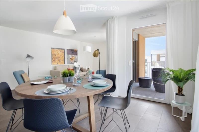 Modern dining area with a round wooden table, six chairs, and a sofa in the background. The room features large windows with white curtains, a pendant light, and a view of a balcony with outdoor seating.
