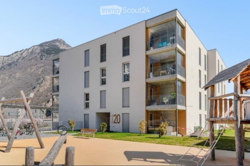 Modern multi-story residential building with large windows and balconies, set against a mountain backdrop. The foreground includes a playground with wooden structures and a bench.