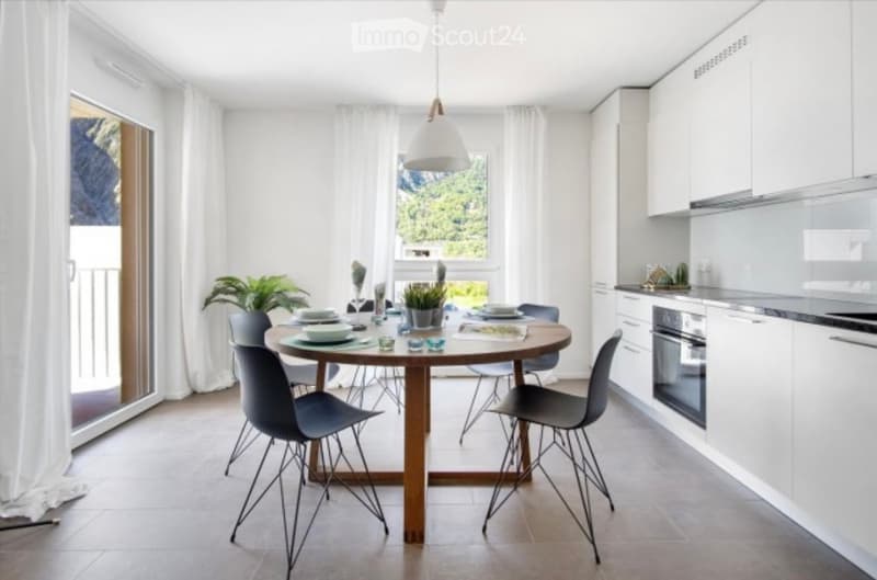 Open-plan kitchen and dining area with white cabinets, a round wooden dining table, modern chairs, and a pendant light. Large windows offer a scenic view.